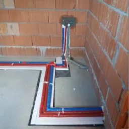 Instalacja wodna i kanalizacyjna w trakcie montażu w surowym wnętrzu, rury czerwone i niebieskie prowadzone wzdłuż ściany z cegły, widoczne przyłącze w ścianie i rura kanalizacyjna.