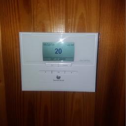 Olesek Instalacje. Projektowanie i wykonywanie instalacji sanitarnych - Biały regulator temperatury Saunier Duval Exacontrol na drewnianej ścianie, wyświetlający temperaturę 20 stopni Celsjusza.