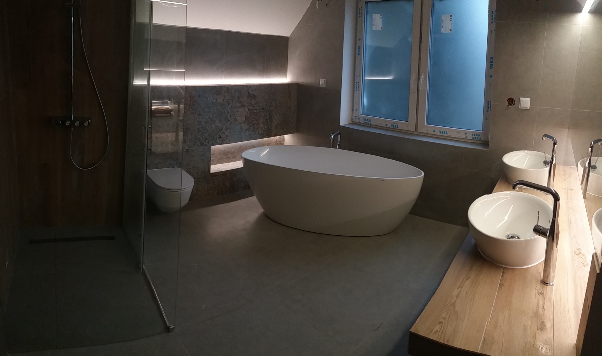 Nowoczesna łazienka z wolnostojącą wanną, kabiną prysznicową typu walk-in, umywalkami nablatowymi na drewnianym blacie i szarą glazurą z podświetleniem LED.