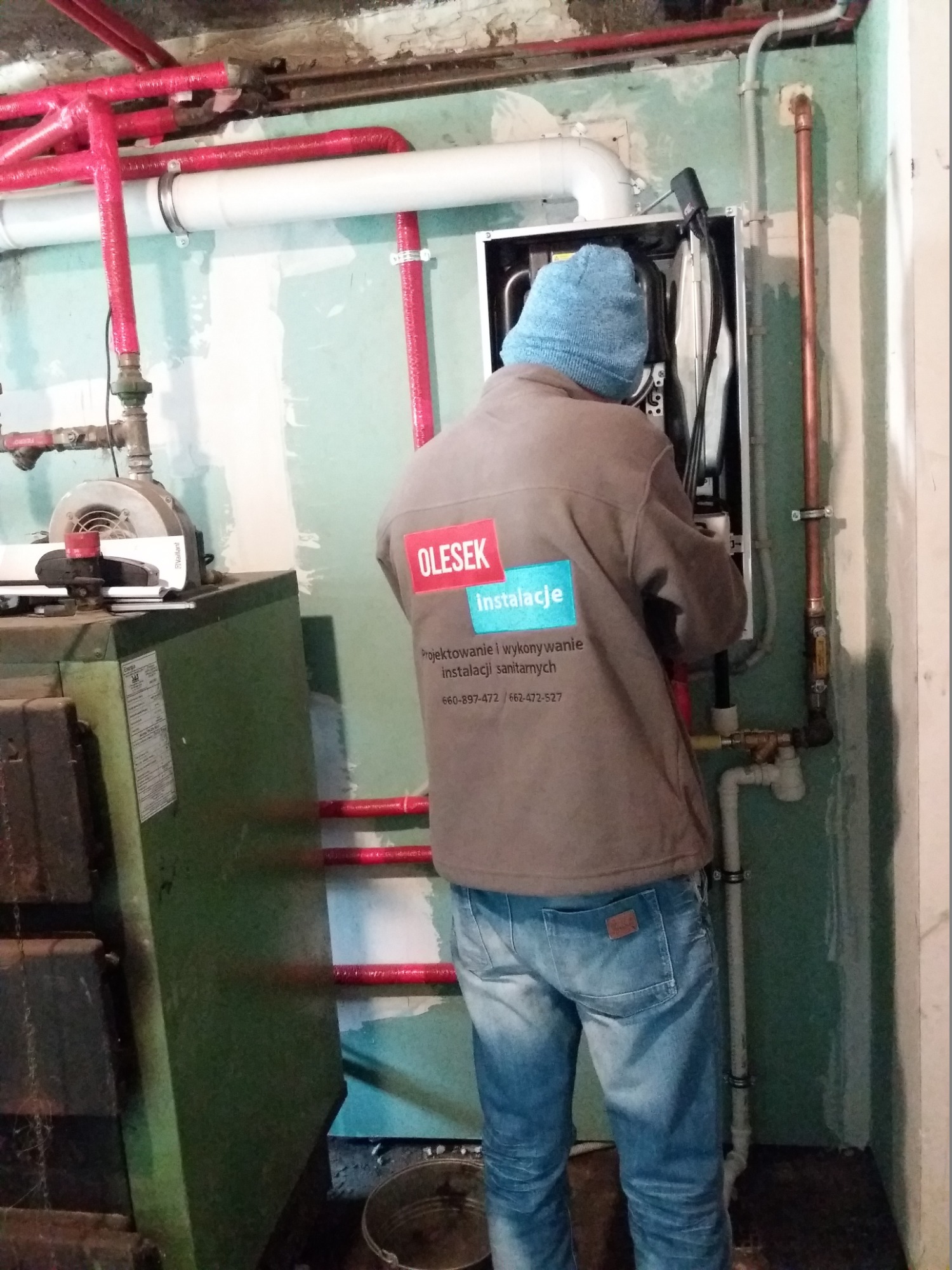 Instalator w niebieskiej czapce w trakcie montażu pieca CO, widoczne rury instalacji grzewczej w izolacji, oraz starszy piec w tle