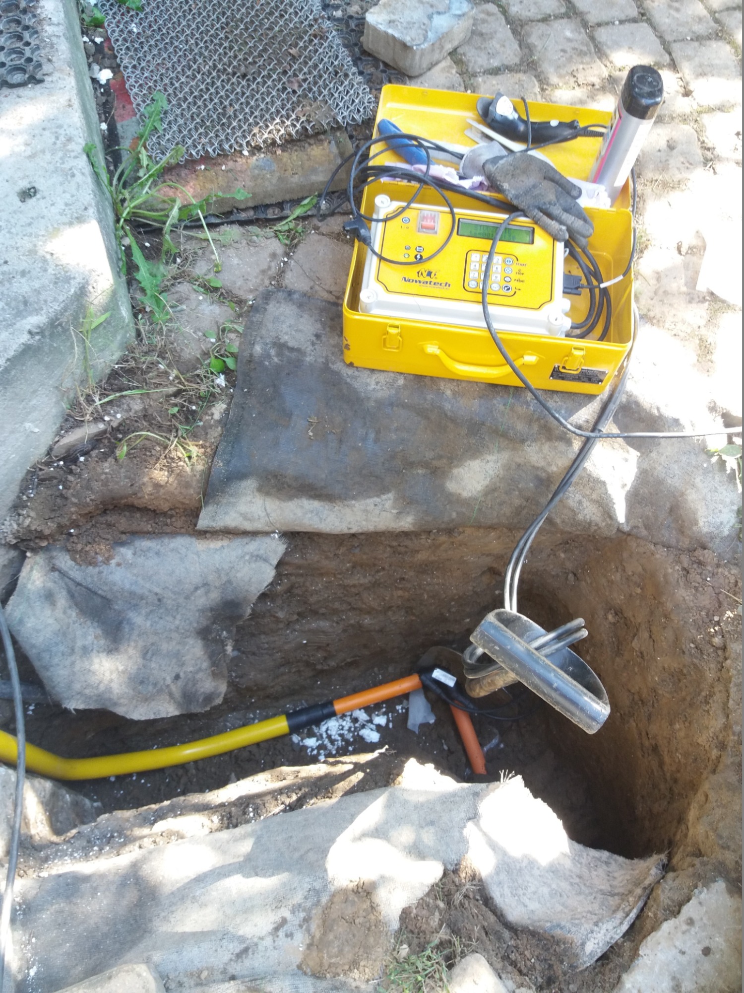 Żółty przewód gazowy w wykopie, narzędzia i detektor nieszczelności Nowatech w żółtej walizce na powierzchni.