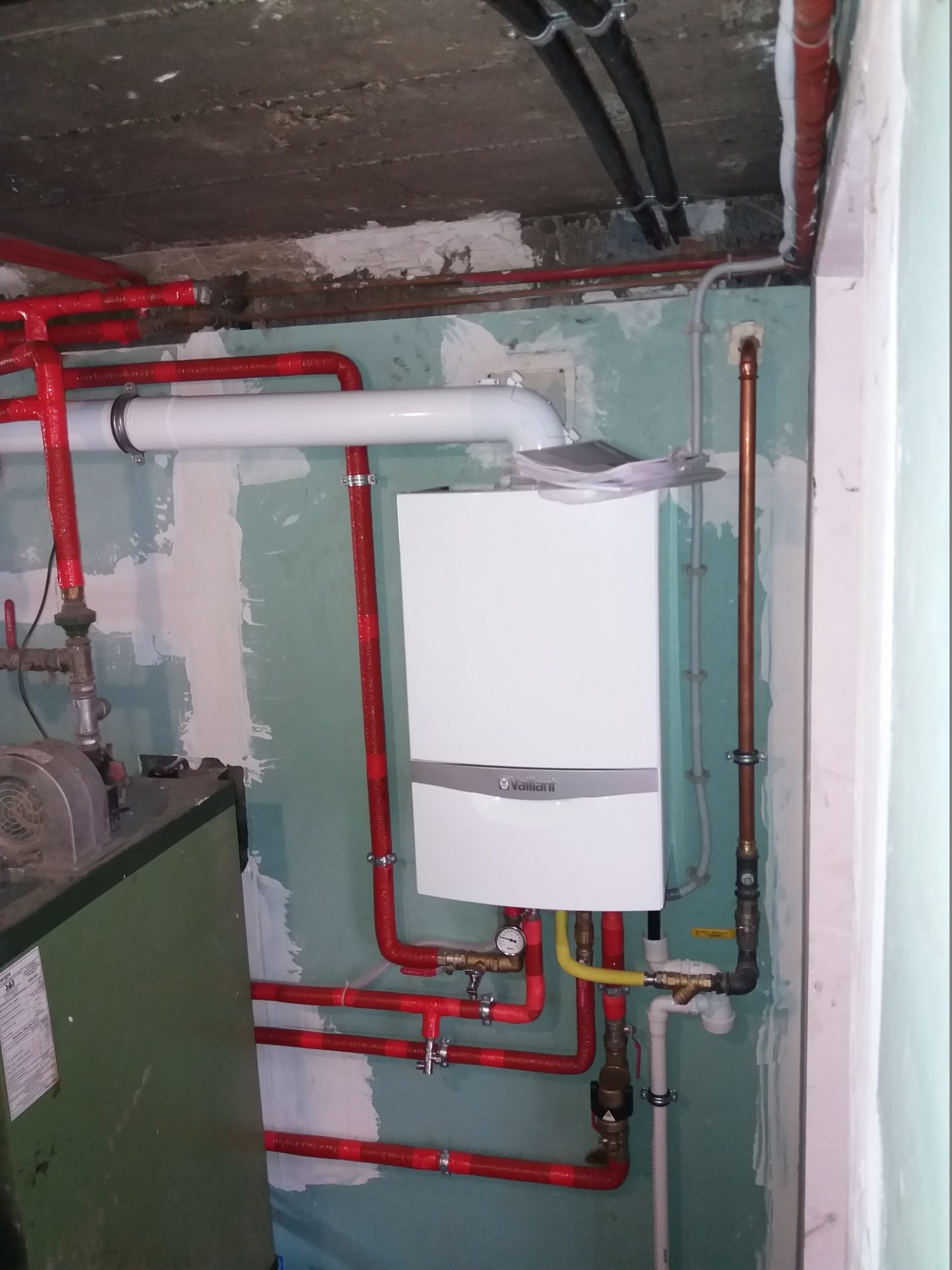 Biały piec gazowy Vaillant zamontowany na ścianie, widoczne rury w izolacji koloru czerwonego, instalacja hydrauliczna oraz stary piec w tle, surowy stan wykończenia pomieszczenia.