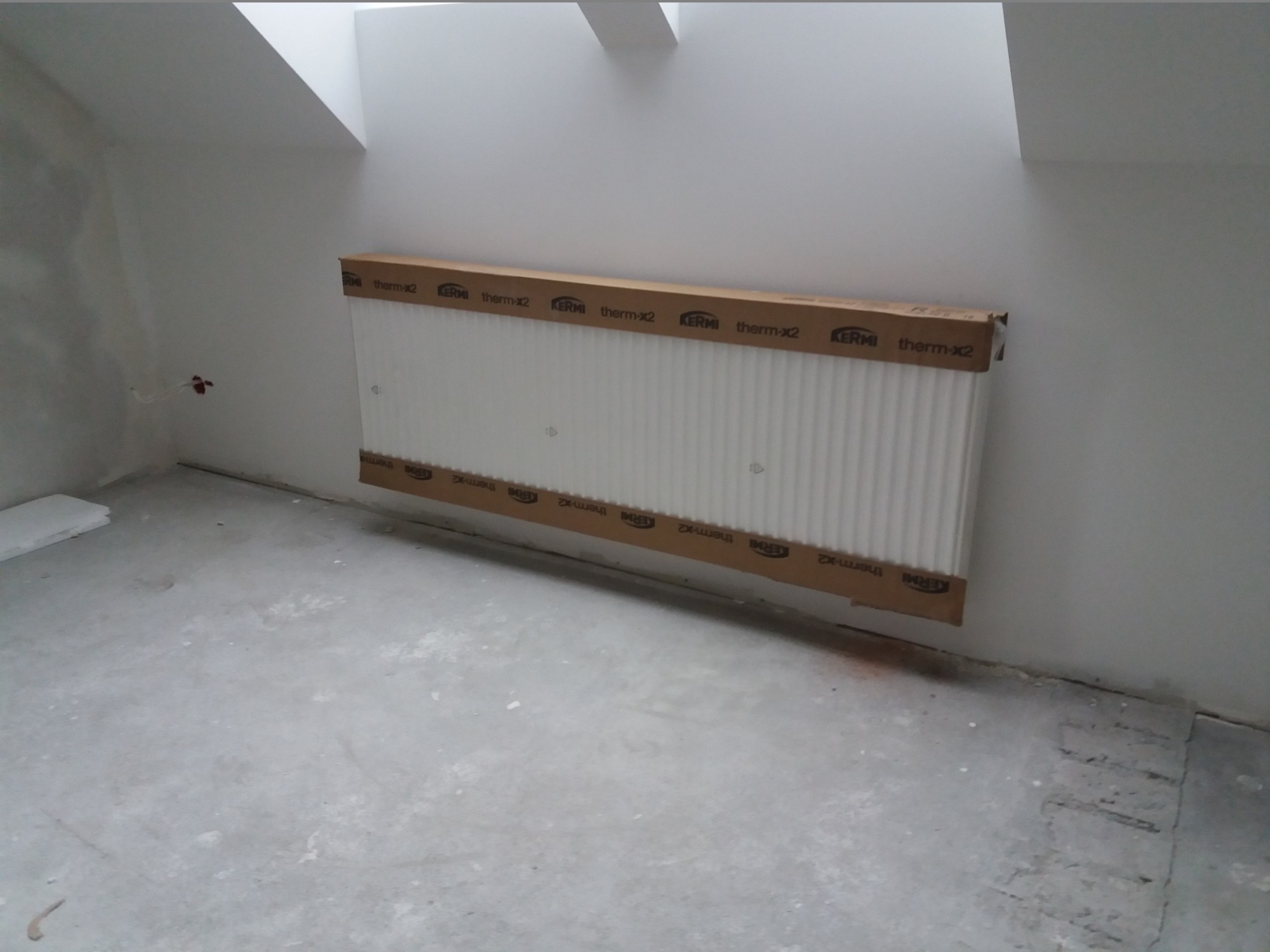 Nowy grzejnik KERMI therm-x2 w opakowaniu kartonowym, przygotowany do montażu w surowym wnętrzu poddasza.