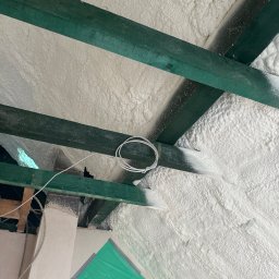 FIRMA HANDLOWO USŁUGOWA Adam Błaszczok - Poddasze z zielonymi belkami i białą pianką izolacyjną, widok z dołu. Fragment instalacji elektrycznej. Folia ochronna w rogu kadru.