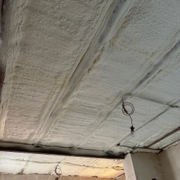 FIRMA HANDLOWO USŁUGOWA Adam Błaszczok - Poddasze ocieplone pianką PUR, widoczne drewniane belki konstrukcyjne i fragment instalacji elektrycznej z żarówką. Aplikacja pianki równomierna, jasna barwa.