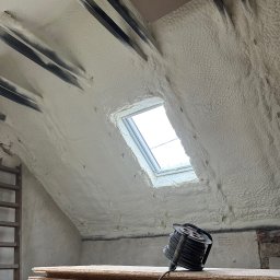 FIRMA HANDLOWO USŁUGOWA Adam Błaszczok - Poddasze po ociepleniu pianką PUR, widoczne okno dachowe, drewniane elementy konstrukcyjne i zwój kabla na deskach. Biała, porowata struktura pianki pokrywa skośne ściany.