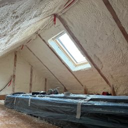 FIRMA HANDLOWO USŁUGOWA Adam Błaszczok - Poddasze po natrysku pianki PUR, widoczne drewniane elementy konstrukcyjne i okno dachowe. Na podłodze zabezpieczone materiały budowlane i narzędzia.