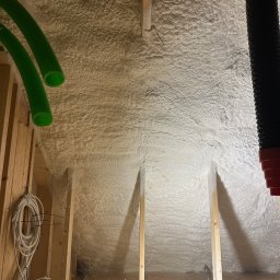 FIRMA HANDLOWO USŁUGOWA Adam Błaszczok - Poddasze ocieplone pianką PUR z widocznymi elementami konstrukcji drewnianej, przewodami wentylacyjnymi i kablami elektrycznymi. Widok z dołu na ocieploną powierzchnię.