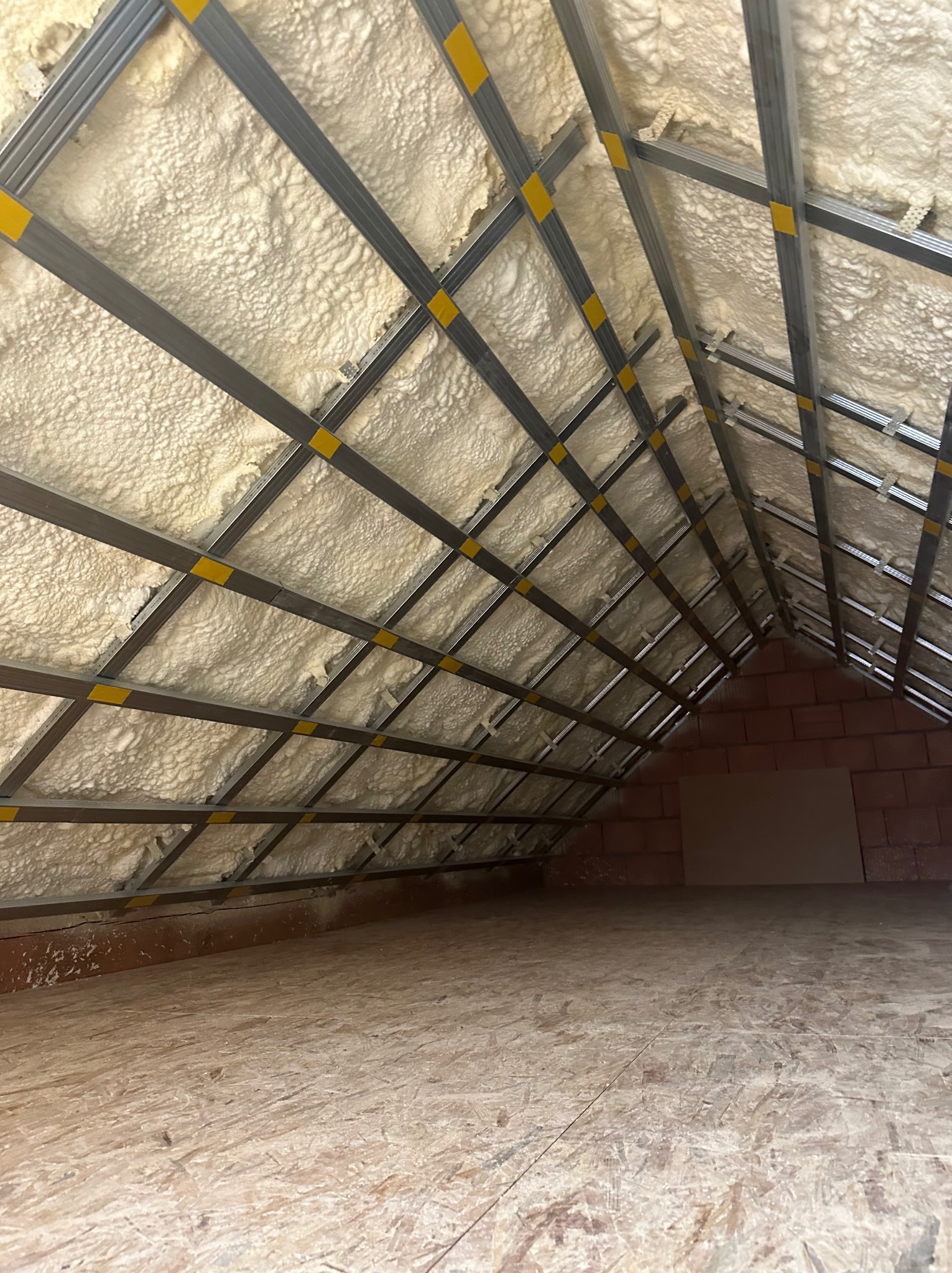 Poddasze ocieplone pianką PUR, widoczne metalowe profile konstrukcyjne z żółtymi taśmami, ściana z czerwonej cegły w tle, podłoga z płyt OSB.