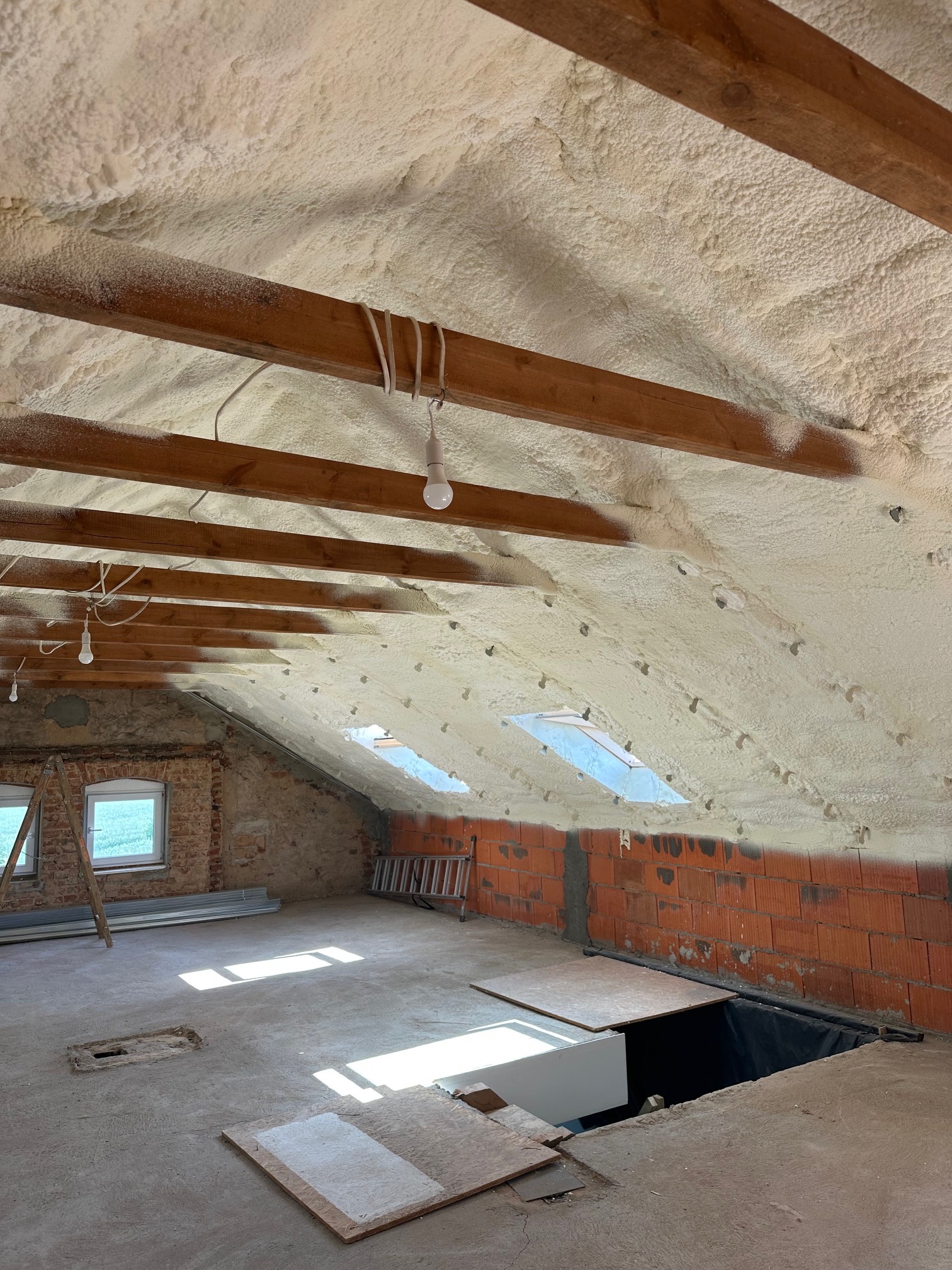 Poddasze po ociepleniu pianką PUR: widoczne drewniane belki i warstwa pianki, okna dachowe. Część ścian z cegły, podłoga betonowa, drabina.
