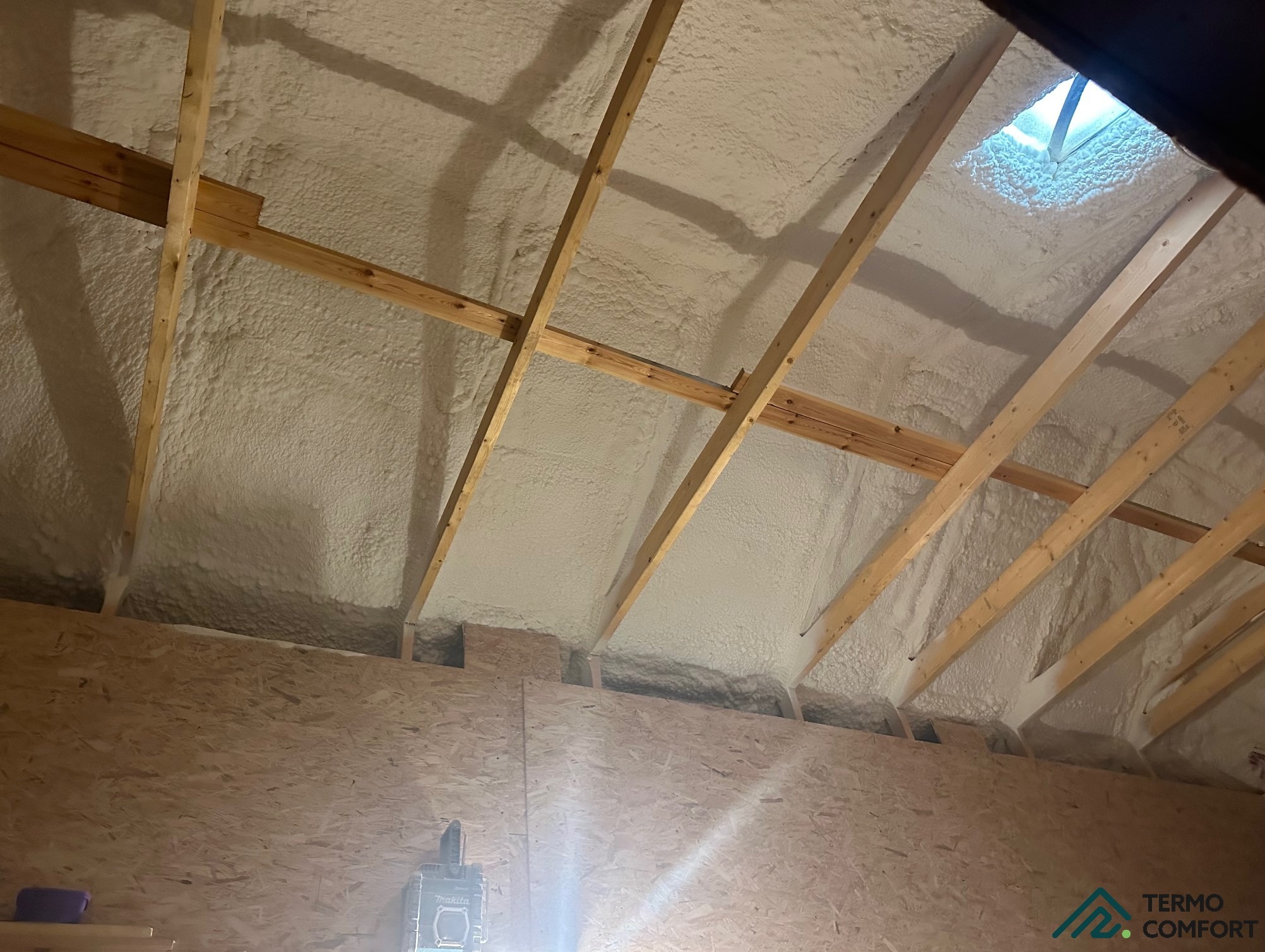 Poddasze ocieplone pianką PUR, widoczne drewniane krokwie i skos dachu z oknem. Na dole ściana z płyty OSB i lampa budowlana. Biała, równomierna warstwa izolacji.
