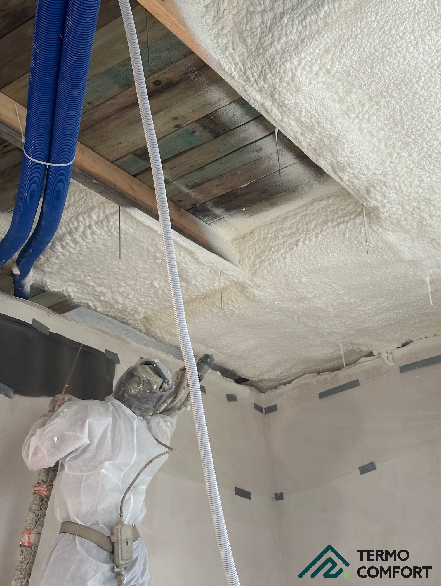 Aplikacja pianki PUR na poddaszu przez pracownika w kombinezonie ochronnym. Widoczne niebieskie węże i drewniana konstrukcja dachu.