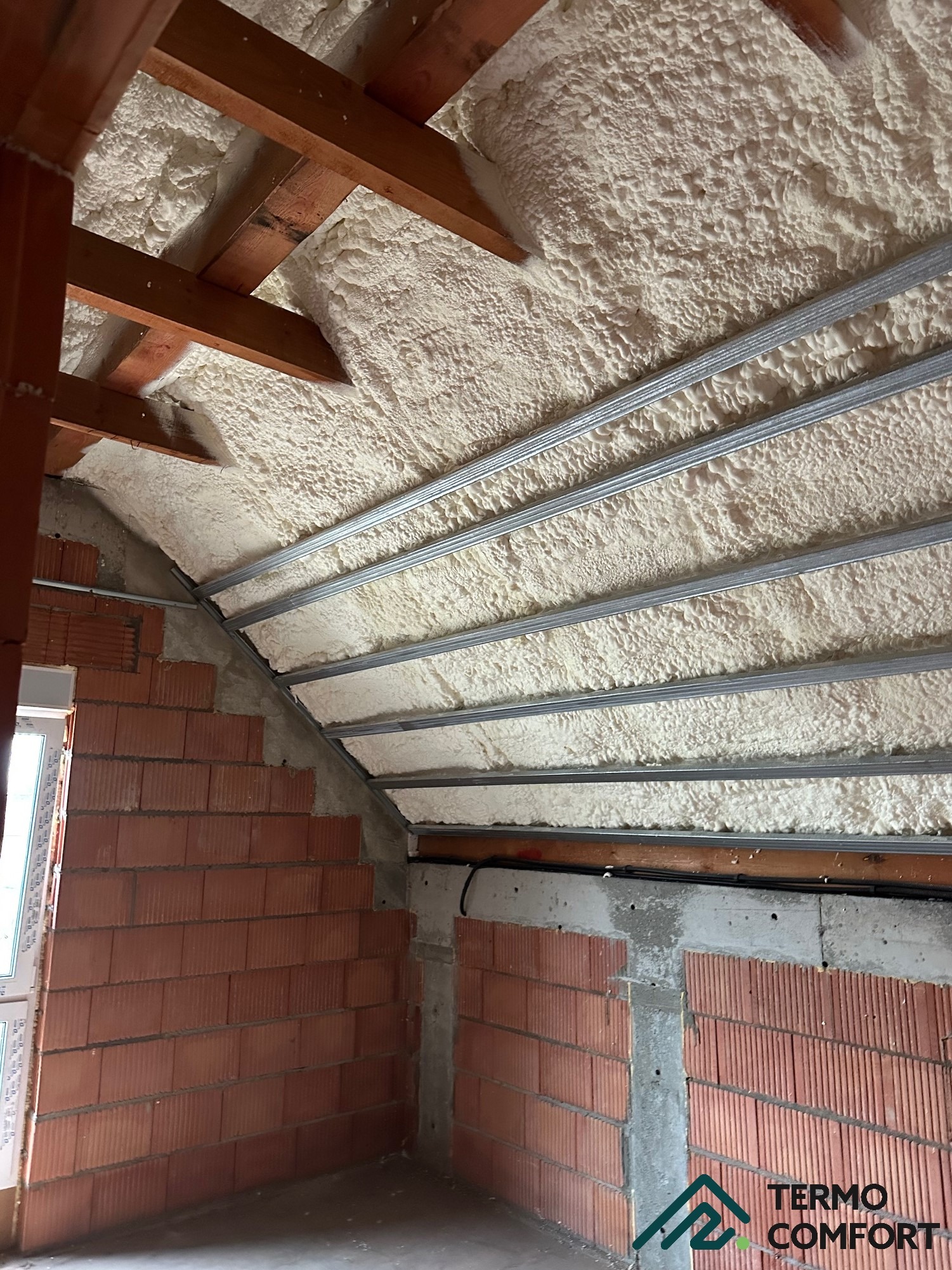 Poddasze ocieplone pianką PUR z widocznymi krokwiami i metalowymi profilami. Ściany z czerwonej cegły dziurawki. Oświetlenie naturalne.