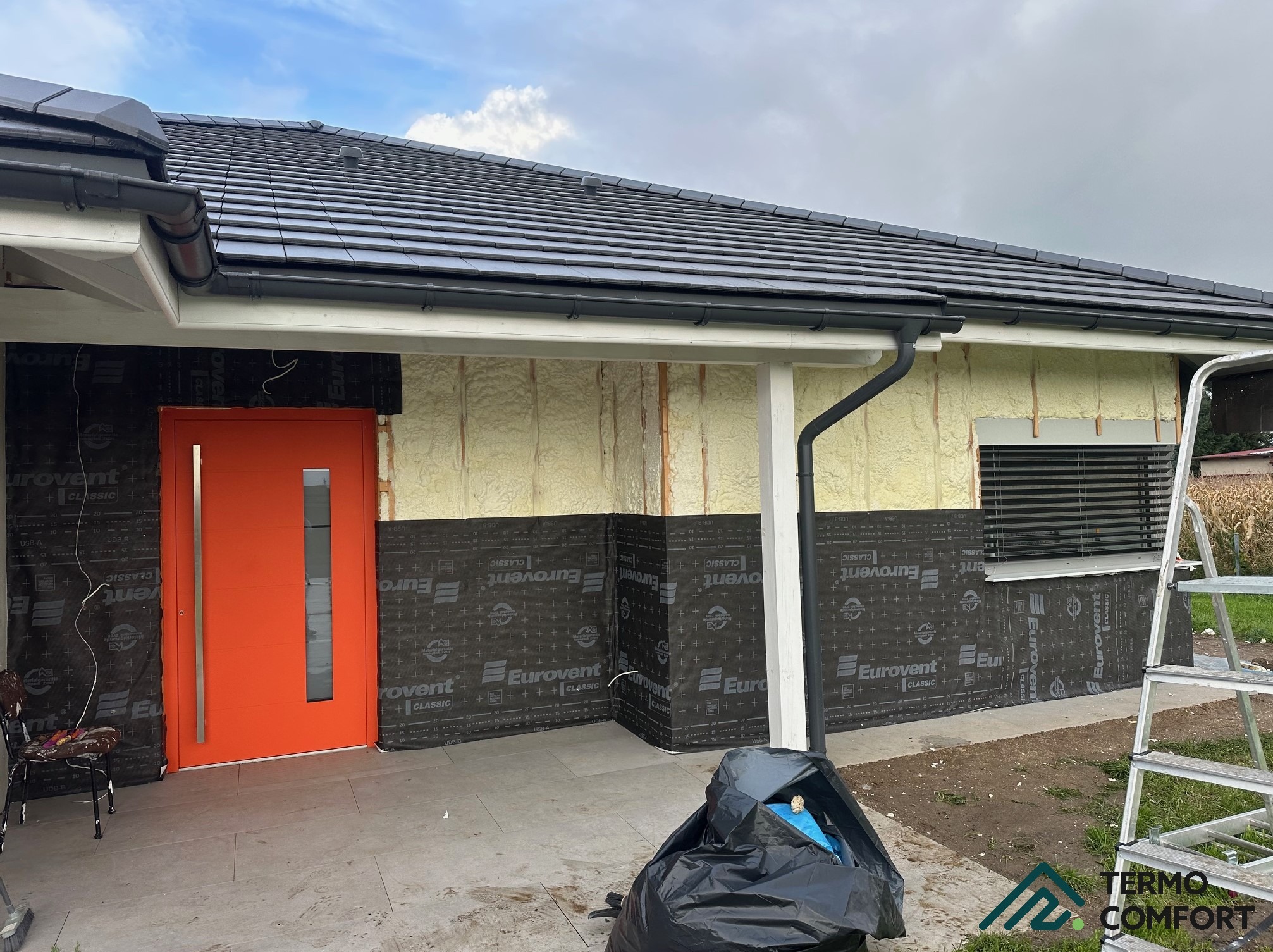 Dom w trakcie ocieplania: pianka PUR widoczna na ścianach, folia Eurovent, pomarańczowe drzwi wejściowe. Etap prac przed pełnym wykończeniem elewacji.