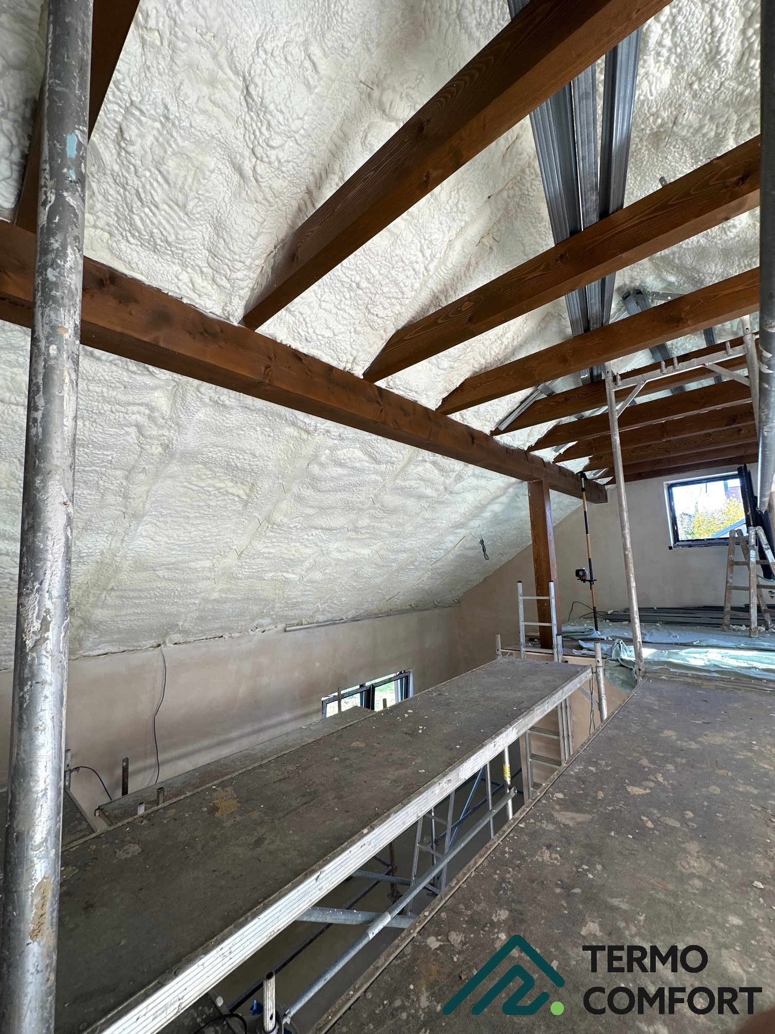Poddasze ocieplone pianką PUR, widoczne drewniane belki stropowe i metalowe profile. Prace wykonane z rusztowania, w tle okno. Izolacja natryskowa.