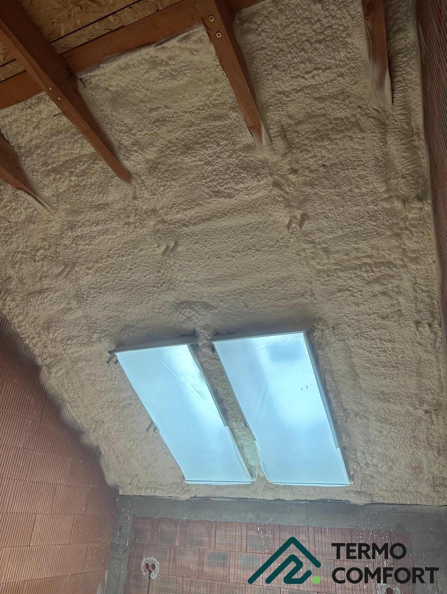 Poddasze ocieplone pianką PUR, widoczne dwa okna dachowe z folią ochronną, drewniane elementy konstrukcyjne i ceglana ściana. Aplikacja pianki równomierna, bez prześwitów.