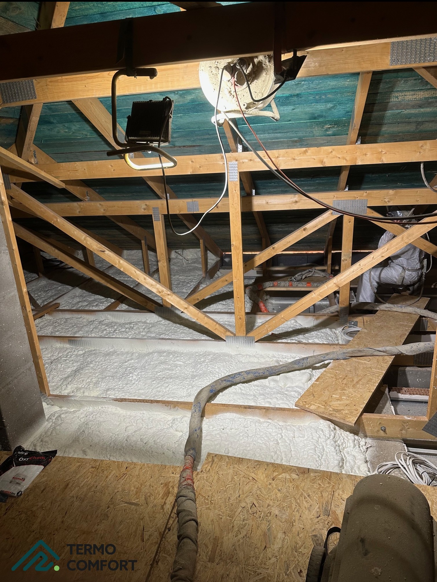 Poddasze z drewnianą konstrukcją, częściowo wypełnione białą pianką izolacyjną. Widoczny wąż do natrysku pianki i pracownik w kombinezonie ochronnym.