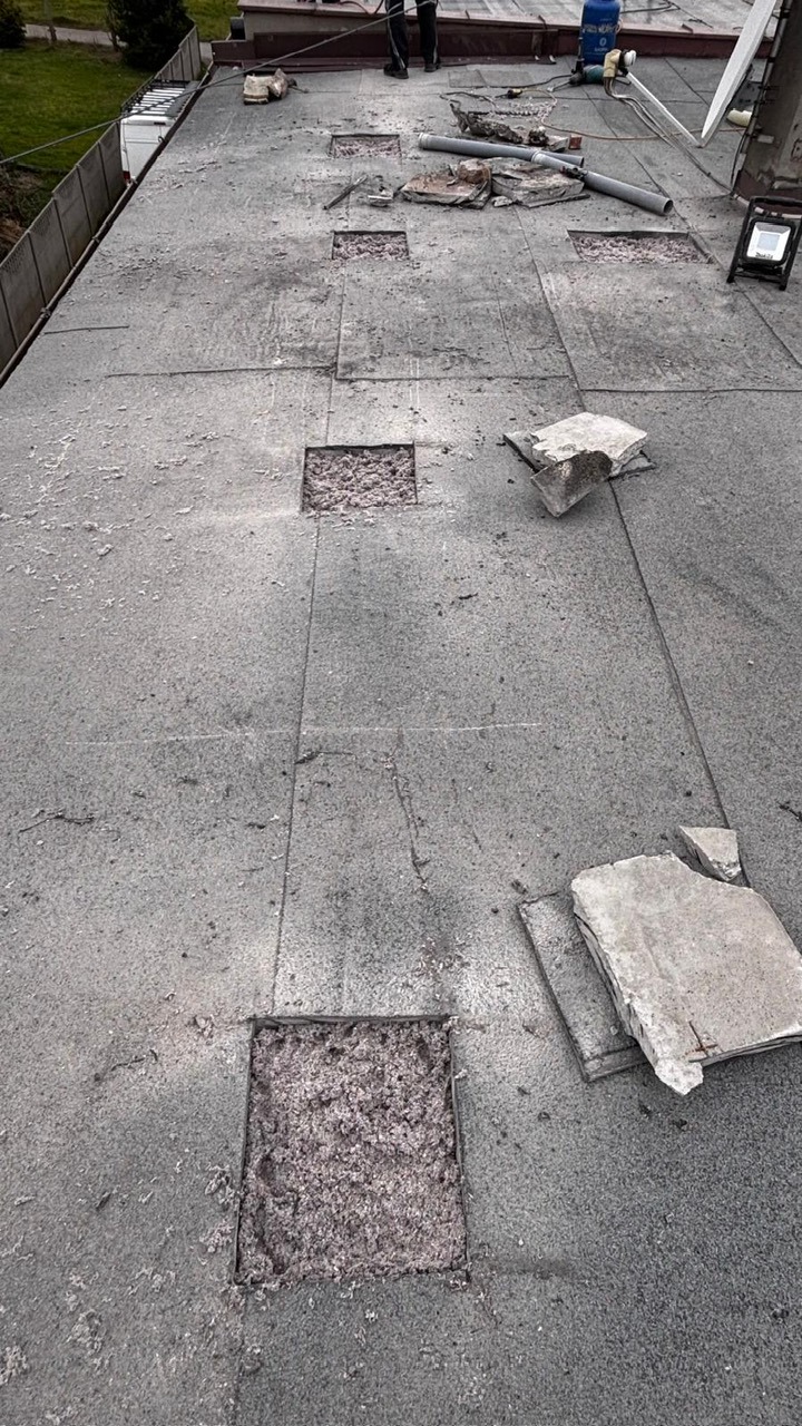 Płaski dach z wyciętymi fragmentami, ukazującymi warstwę izolacyjną. Widoczne narzędzia i materiały budowlane, sugerujące etap przygotowania do ocieplenia.