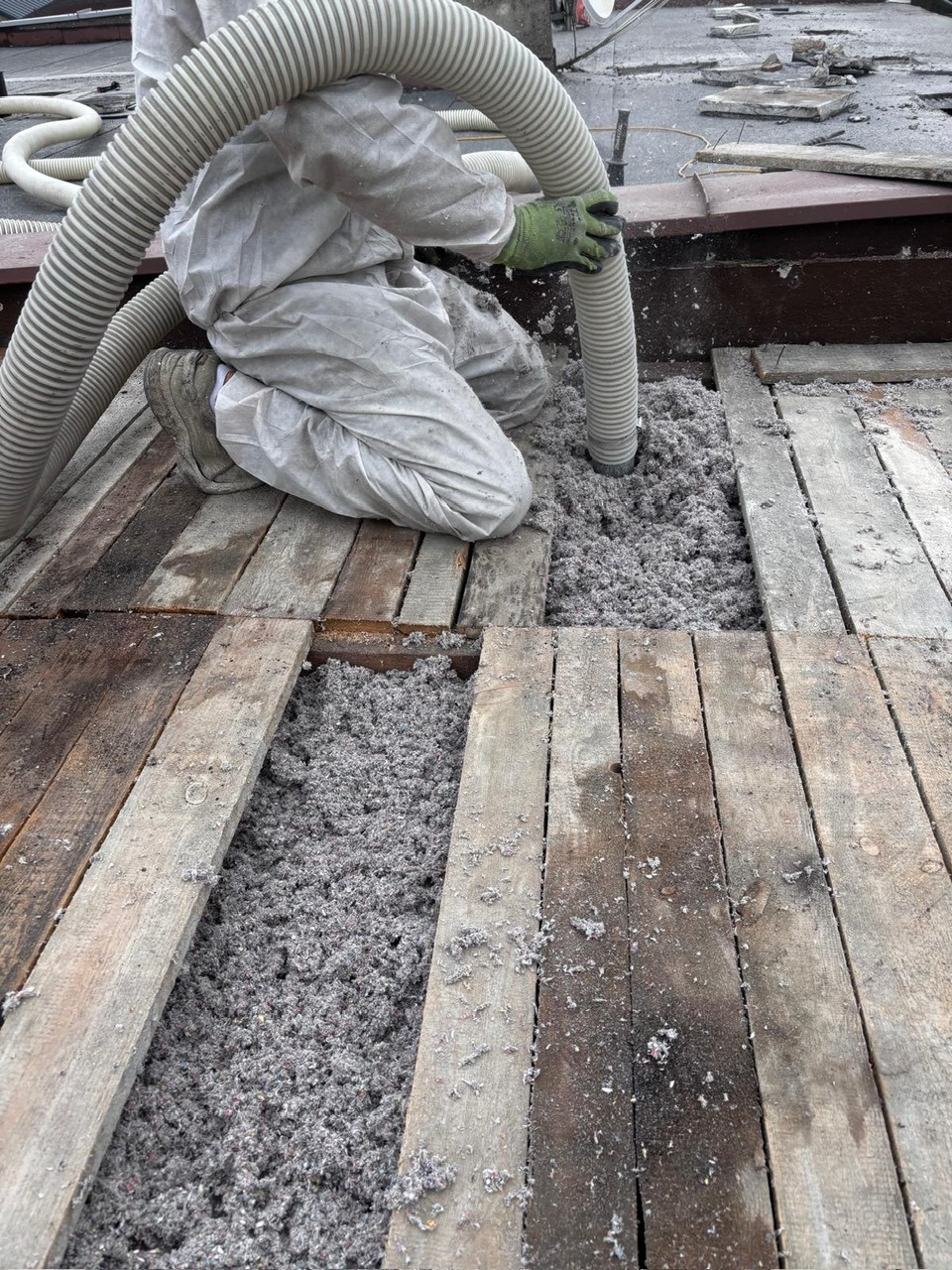 Pracownik w kombinezonie aplikuje szarą izolację na drewnianą konstrukcję poddasza za pomocą węża. Widoczne fragmenty podłogi i izolowany obszar.