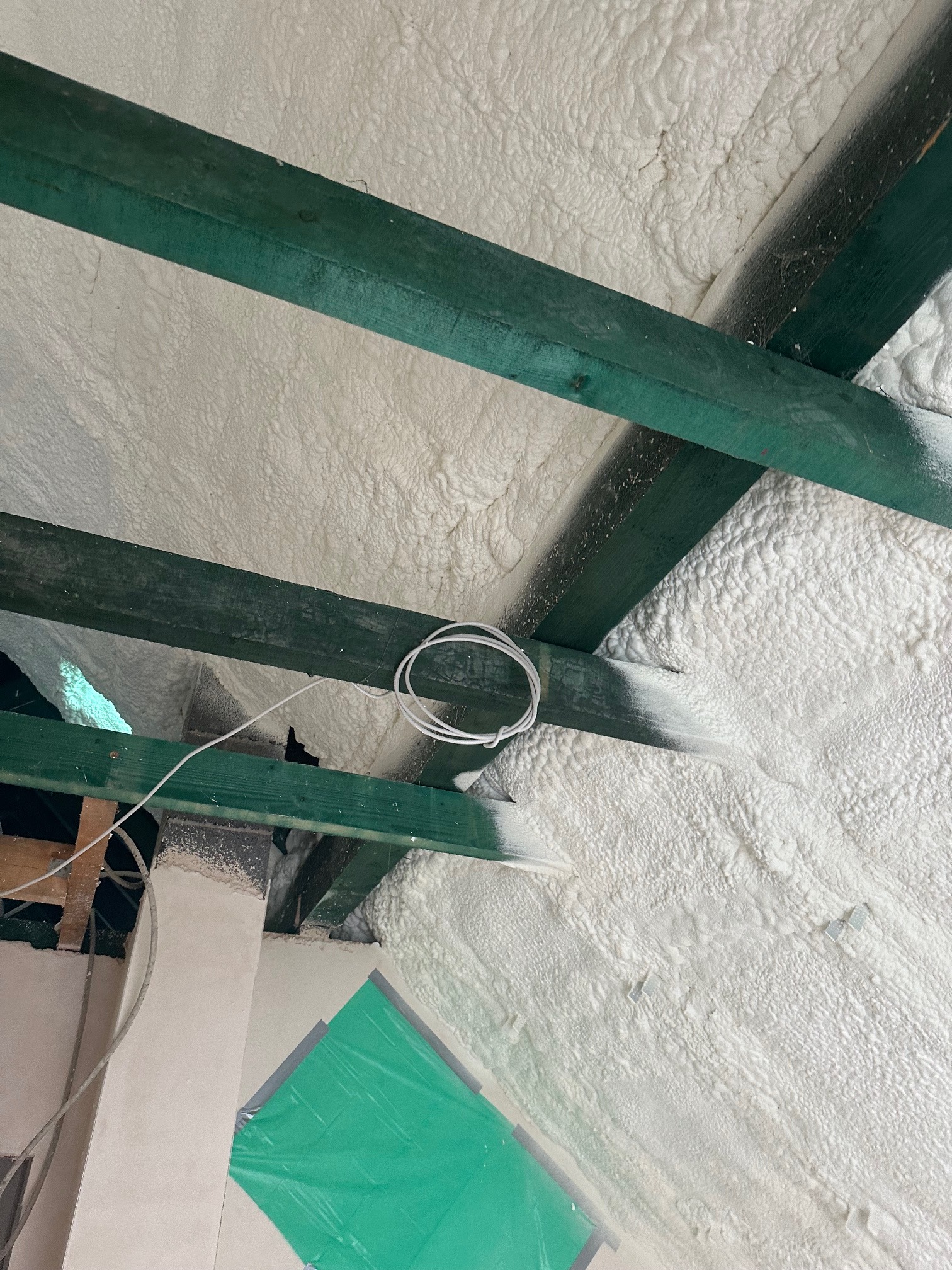 Poddasze z zielonymi belkami i białą pianką izolacyjną, widok z dołu. Fragment instalacji elektrycznej. Folia ochronna w rogu kadru.