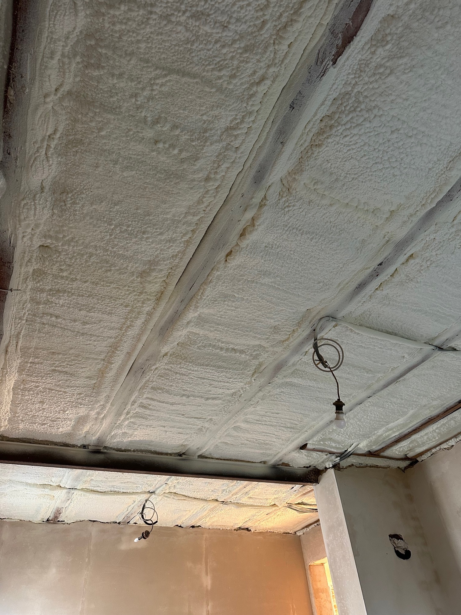 Poddasze ocieplone pianką PUR, widoczne drewniane belki konstrukcyjne i fragment instalacji elektrycznej z żarówką. Aplikacja pianki równomierna, jasna barwa.