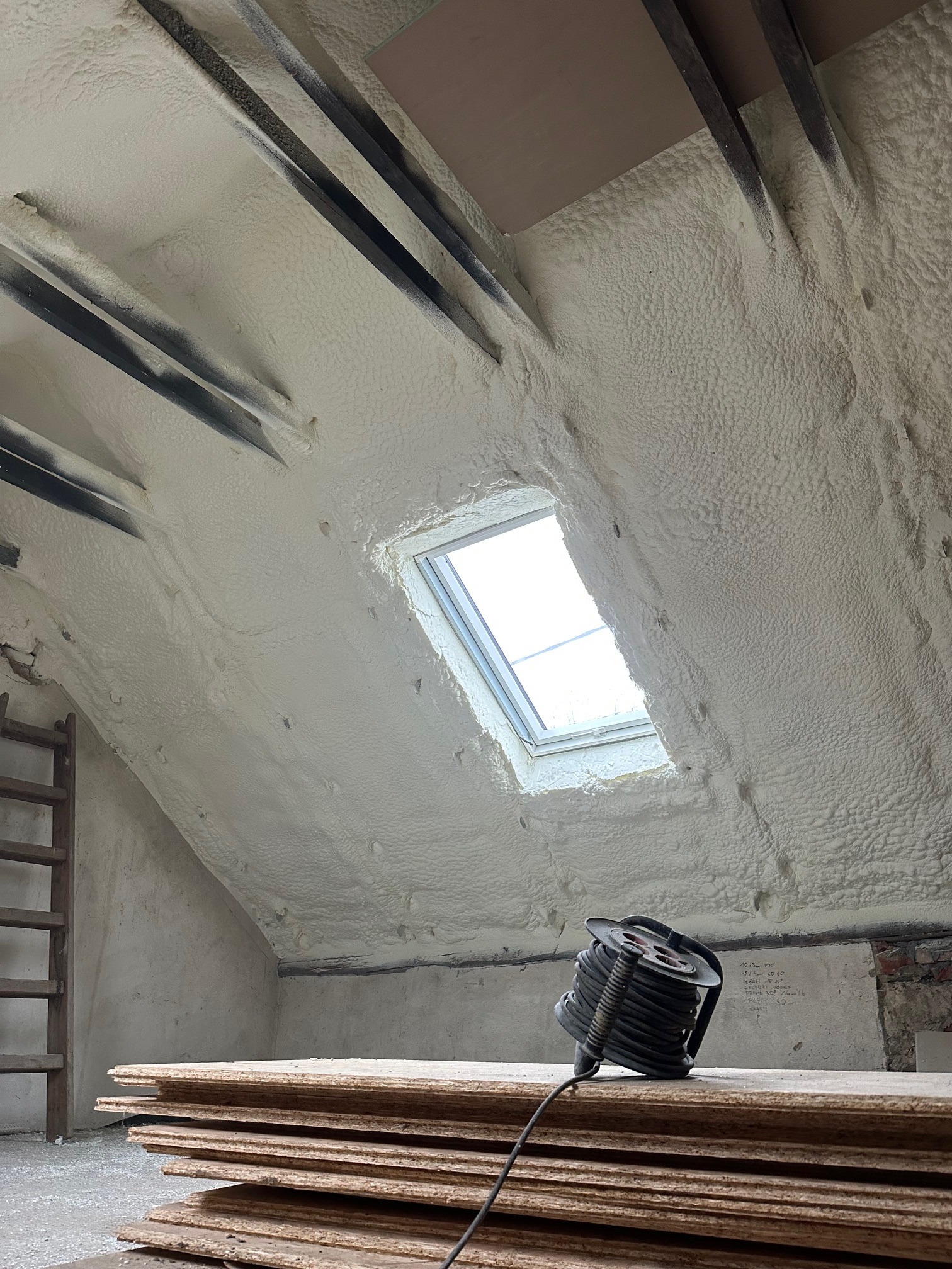 Poddasze po ociepleniu pianką PUR, widoczne okno dachowe, drewniane elementy konstrukcyjne i zwój kabla na deskach. Biała, porowata struktura pianki pokrywa skośne ściany.