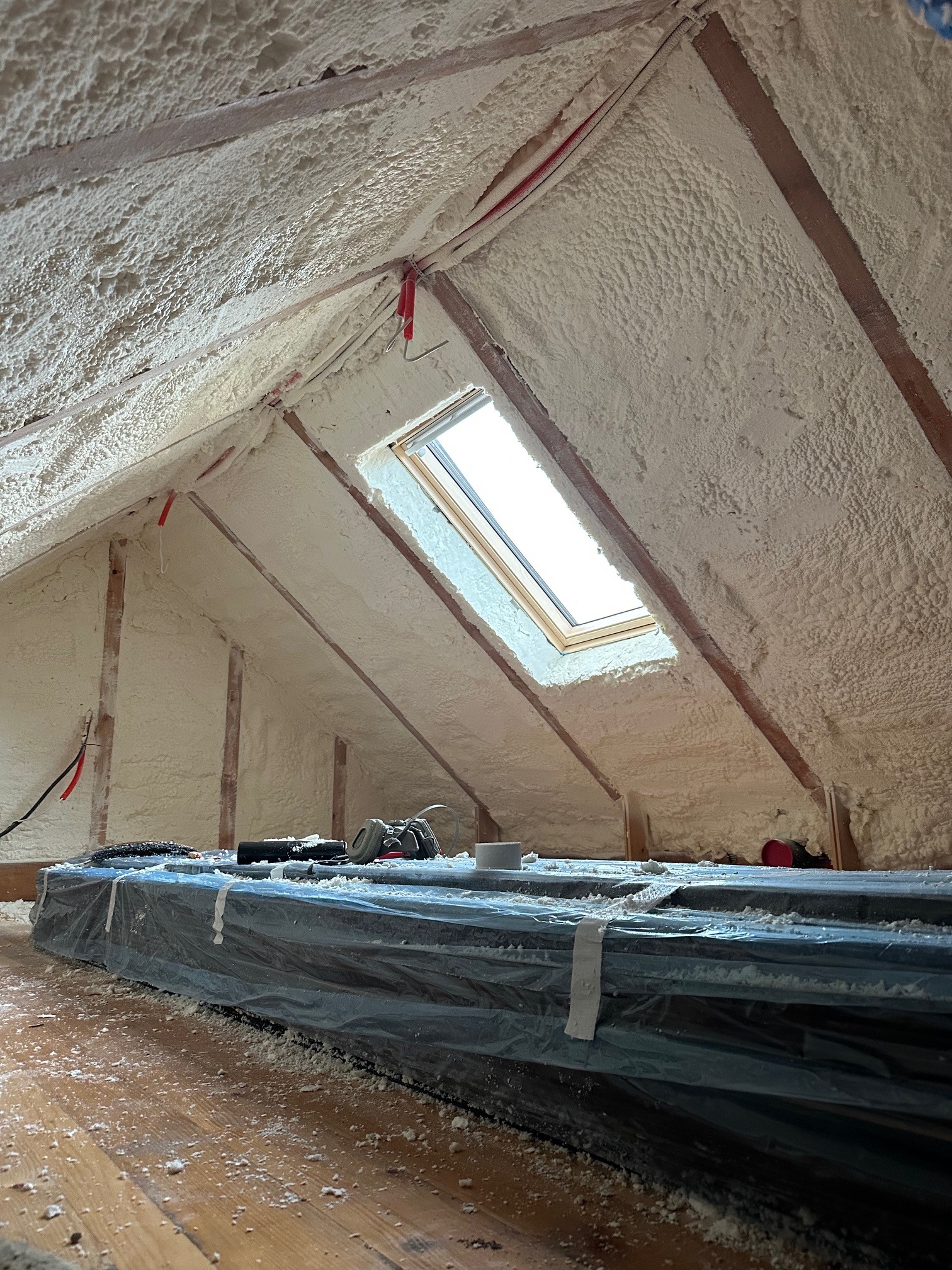 Poddasze po natrysku pianki PUR, widoczne drewniane elementy konstrukcyjne i okno dachowe. Na podłodze zabezpieczone materiały budowlane i narzędzia.