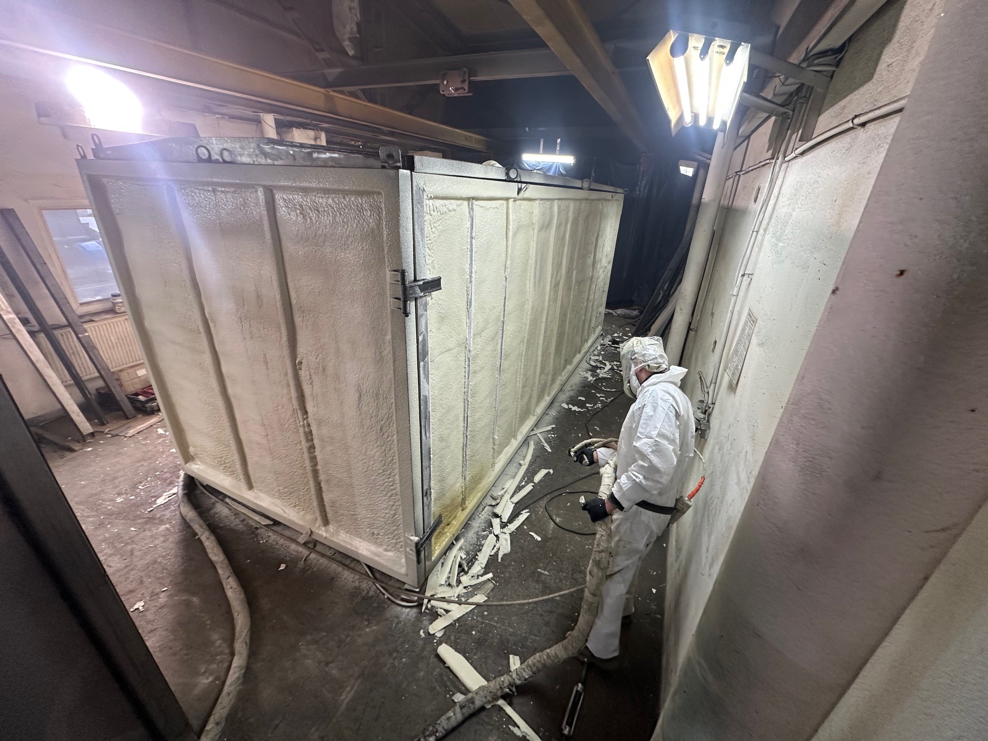 Pracownik w kombinezonie ochronnym aplikuje piankę PUR na metalową konstrukcję. Widoczne węże, resztki pianki na podłodze i oświetlenie przemysłowe.