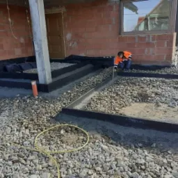 Przygotowanie podłoża pod taras: pracownik wyrównuje podsypkę z kamienia w obramowaniu z czarnego tworzywa, widoczny fragment niedokończonego budynku z cegły.