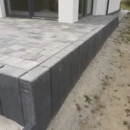 Wykończony taras z szarej kostki brukowej, obrzeże z pionowo ułożonych, ciemnych kostek. Widok na elewację budynku i fragment ogrodu.