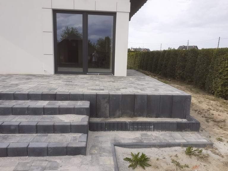 Ułożona kostka brukowa w Kazimierzy Wielkiej tworząca schody i taras przed domem z szarymi panelami i dużymi oknami, obok zielony żywopłot.