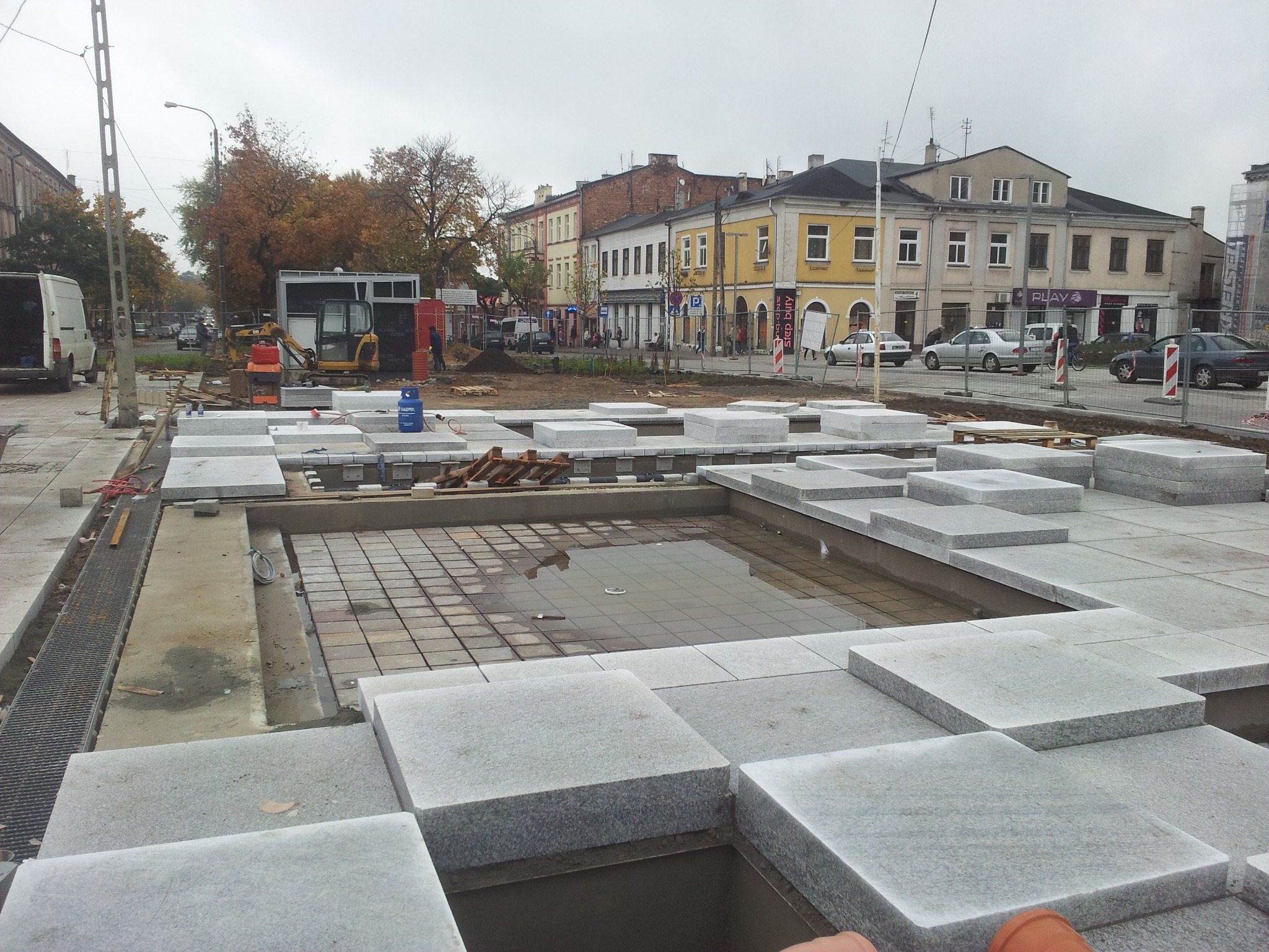 Układanie kostki granitowej na placu miejskim z widocznymi elementami infrastruktury i mini-koparką w tle.