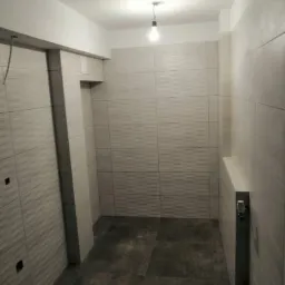 Wykończona łazienka z szarymi płytkami ściennymi o falistej strukturze i ciemnymi płytkami podłogowymi, widoczny grzejnik drabinkowy, surowa żarówka zwisająca z sufitu.