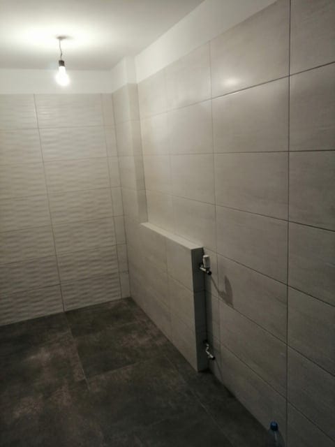 Wykończona łazienka z jasnymi, kwadratowymi płytkami ściennymi, ciemną podłogą z płytek i grzejnikiem drabinkowym z widocznymi zaworami.