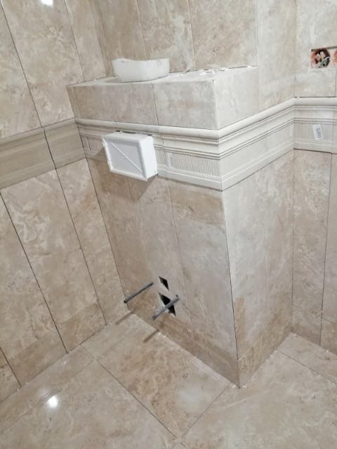 Ściana i podłoga w łazience wyłożone beżowymi płytkami, widoczne wystające rury i białe elementy instalacji.