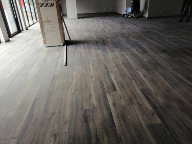 Ułożona podłoga z płytek imitujących drewno w nowym, przestronnym pomieszczeniu biurowym, widoczny karton z napisem OKOCIM.