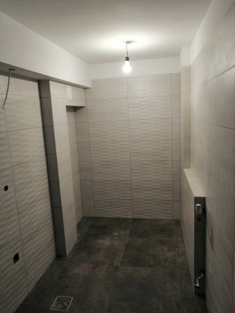 Wykończona łazienka z szarymi płytkami ściennymi o falistej strukturze i ciemnymi płytkami podłogowymi, widoczny grzejnik drabinkowy, surowa żarówka zwisająca z sufitu.