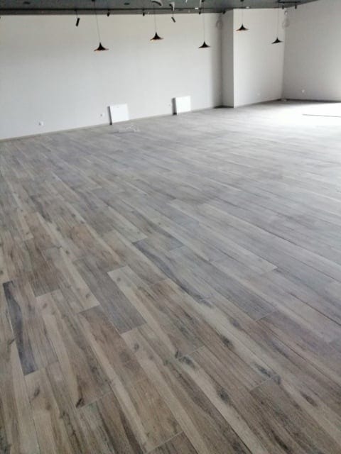 Ułożona podłoga z płytek imitujących drewno w pustym, jasnym pomieszczeniu biurowym z białymi ścianami i wiszącymi lampami.