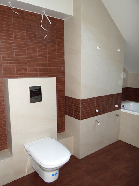 Nowoczesna łazienka z brązowo-beżowymi płytkami, wisząca toaleta z czarnym przyciskiem spłukiwania, widoczne niedokończone kable elektryczne wystające z sufitu.