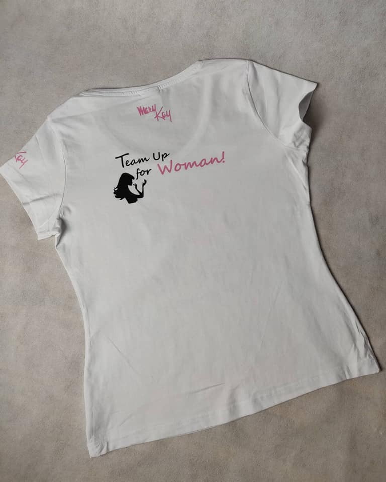 Biała koszulka damska z nadrukiem 'Team Up for Woman!' i logo Mary Kay, leżąca na szarym tle.