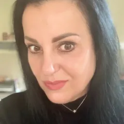 Portret kobiety z ciemnymi włosami, delikatnym makijażem i srebrnym naszyjnikiem z małą zawieszką na jasnym tle.