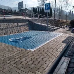 Oznakowane, niebieskie miejsce parkingowe dla osób niepełnosprawnych, wykonane z kostki brukowej, z widocznymi schodami z metalową barierką i znakiem pionowym oznaczającym parking...