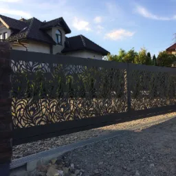 Nowoczesna brama wjazdowa przesuwna z laserowo wycinanym wzorem roślinnym, zamontowana na posesji z domem jednorodzinnym, widok z zewnątrz na podjazd z kamiennym kruszywem.