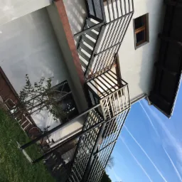 Zewnętrzne schody metalowe z balustradą, prowadzące na balkon w Pietrzykowicach, widoczne na tle trawnika i błękitnego nieba.