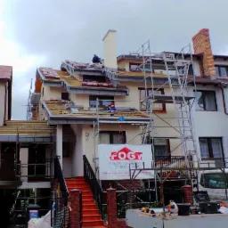 Dom szeregowy w trakcie prac dekarskich, widoczne rusztowania, częściowo pokryty dachówką, na dachu pracownicy, baner firmy budowlanej przed budynkiem.