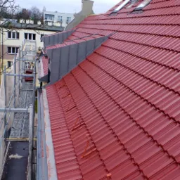 Czerwony dach z dachówki ceramicznej, widok z rusztowania, elementy blaszane w kolorze grafitowym, budynki w tle, pochmurny dzień.