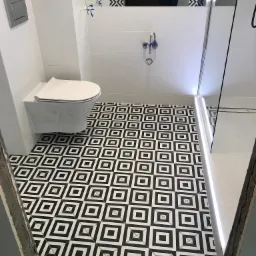 Nowoczesna łazienka z białą armaturą i podwieszaną toaletą, czarno-biała geometryczna podłoga z płytek, widoczny fragment kabiny prysznicowej z oświetleniem LED.