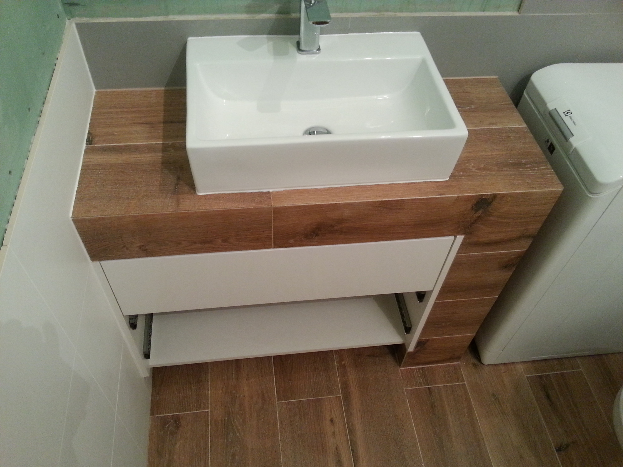 Biała, prostokątna umywalka nablatowa z chromowaną baterią, zamontowana na szafce z frontami w kolorze białym i blatem imitującym drewno, obok pralka Electrolux, całość wykończona płytkami...