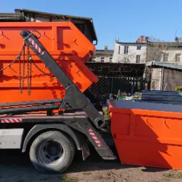 Pomarańczowy kontener na gruz umieszczony na ciężarówce z hydraulicznym ramieniem, drugi kontener stoi obok na ziemi, w tle zabudowania miejskie.