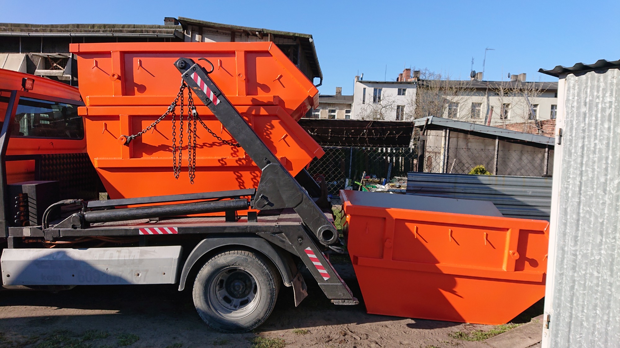 Pomarańczowy kontener na gruz umieszczony na ciężarówce z hydraulicznym ramieniem, drugi kontener stoi obok na ziemi, w tle zabudowania miejskie.